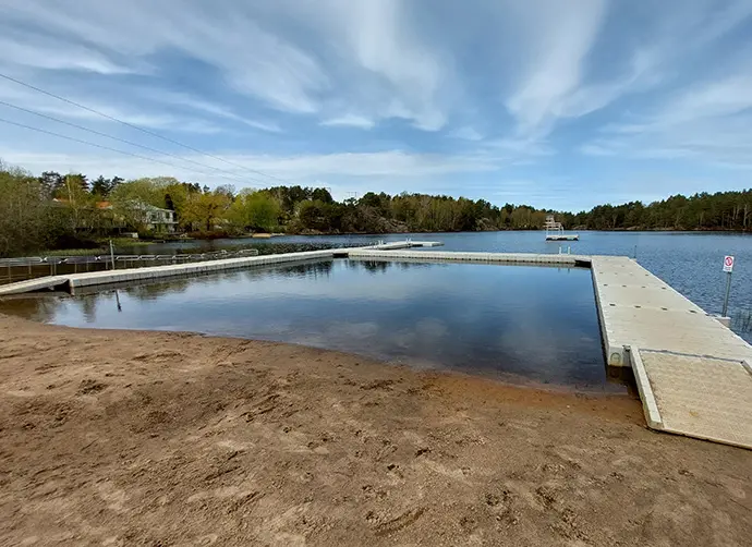 En brygga ligger i en sjö. marken närmast bryggan är täckt av sand. En blå himmel lyser upp området.