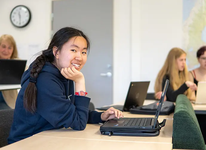 Elev sitter vid laptop i klassrum och ler mot kameran.
