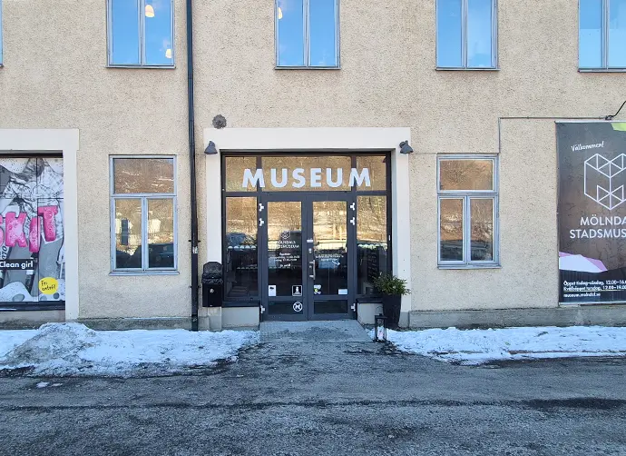 Entré till museet. En ljus fasad och glasportar med texten museum överst. Det är snö på marken.