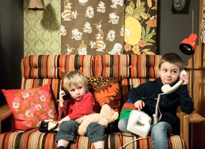 Två mindre barn sitter i en soffa klädd i orange, beige och brunt tyg.  Barnen håller i och låtsas lyssna i varsin telefon.