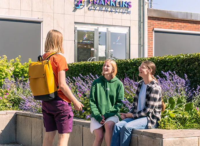 Tre elever sitter utanför skolan och pratar tillsammans vid blomrabatt.