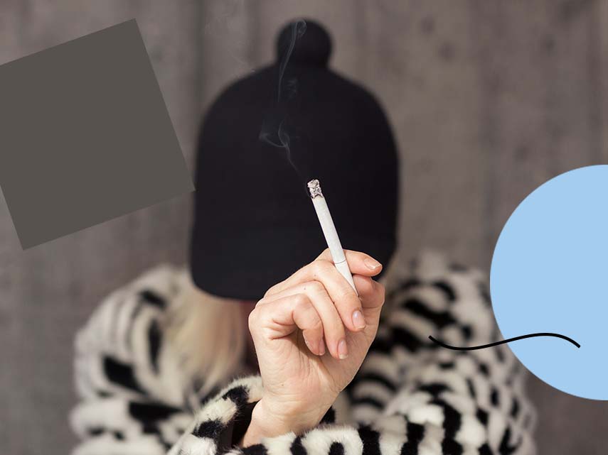 En hand som håller i cigarett. I bakgrunden syns personen som håller i cigaretten, med en mössa över huvudet.