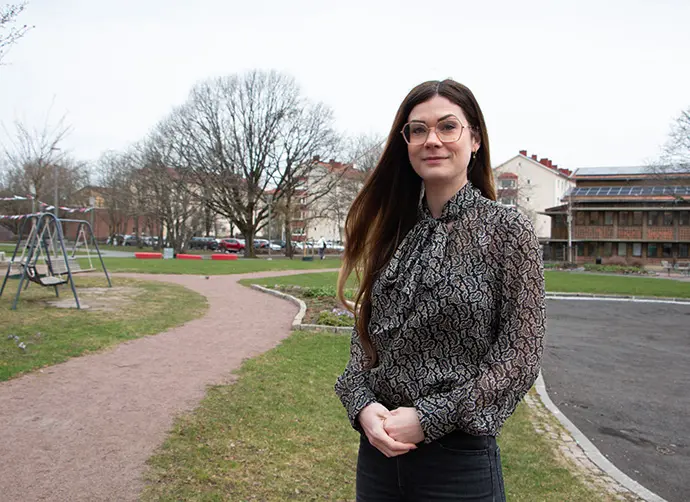 En person står framför en park med en lekplats och ett parkhus i bakgrunden. Personen bär glasögon och en blus med mönster.
