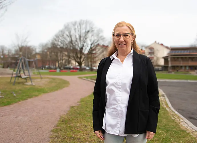 En kvinna med glasögon står framför en park med lekplats och bostadshus. Hon bär en vit skjorta och en svart kavaj.