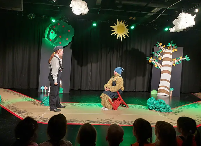 En teaterpjäs där två ungdomar pratar med varandra med. En står i polisuniform och den andra sitter i en björndräkt med träd, moln och en sol i bakgrunden. I publiken sitter flera barn.