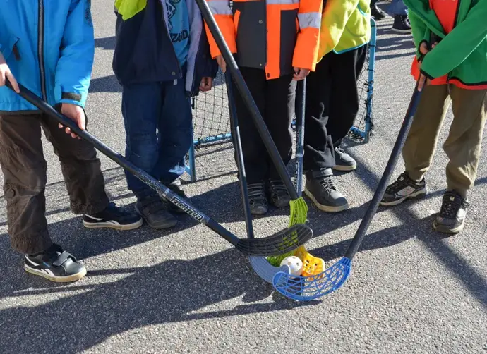 Flera barn står med bandyklubbor vid en asfaltsplan.