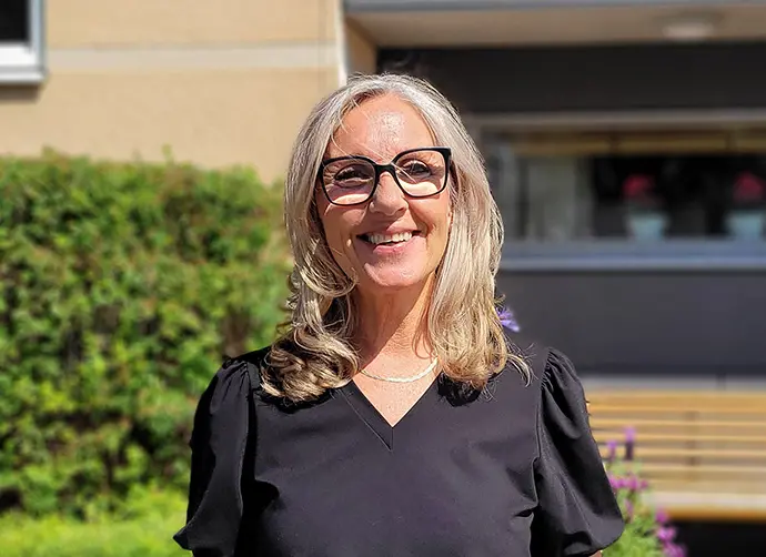 Kristin Algelind, undersköterska, står framför en grön buske och en bänk.
