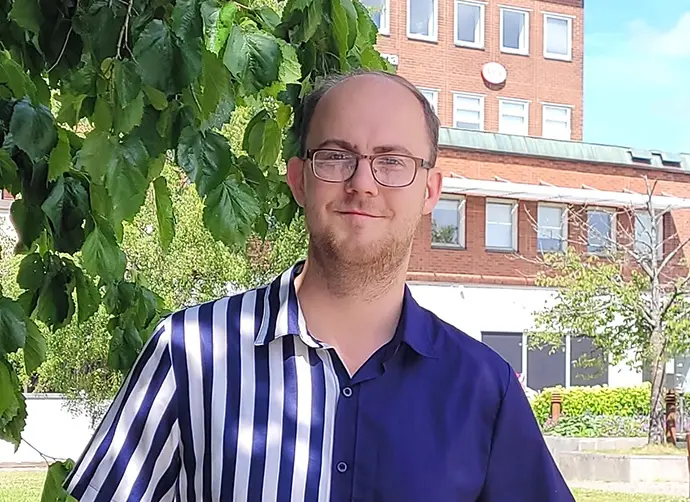 Johannes Öberg, stödpedagog, står under ett träd klädd i blå randig skjorta.