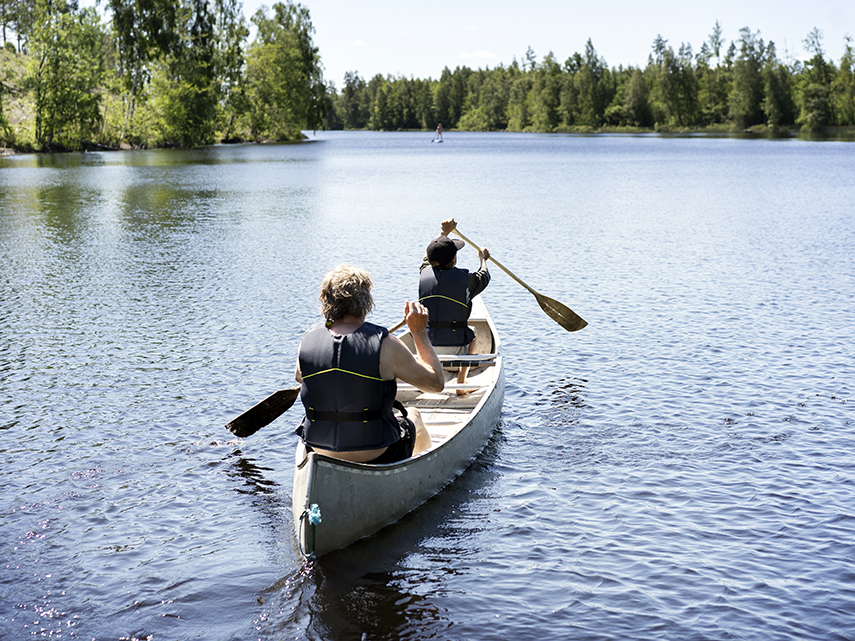 Två personer sitter i en kanot som rör sig ut på en sjö. Längre bort syns en person som står på en Stand-Up paddleboard.