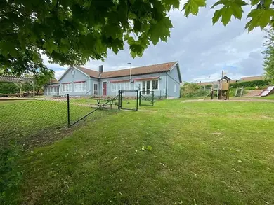 Ett blått hus med en lekplats omgiven av grönt gräs. En lågt staket är synlig i förgrunden, tillsammans med löv från ett träd.