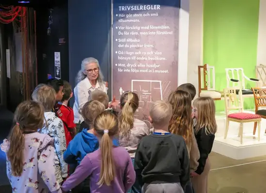 En grupp barn står framför en kvinna och en skylt. Barnen står med ryggen mot kameran. Längst till höger i bild syns flera 1700-talsstolar.