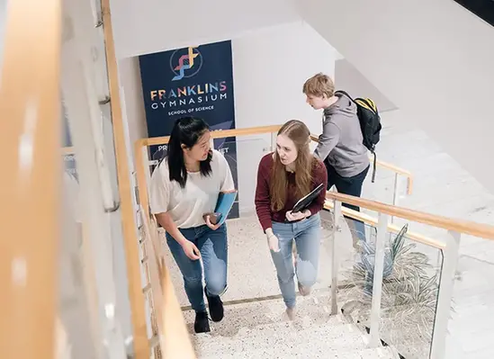 Två elever går uppför en trappa och samtalar medan de bär skolmaterial, med skolans miljö i bakgrunden.