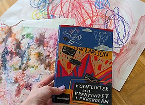 en hand håller en bok över ett bord med barnteckningar