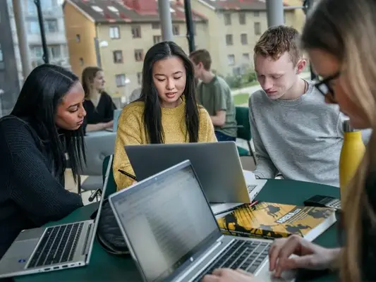 Elever sitter runt ett bord med sina laptops och diskuterar en skoluppgift.
