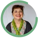 Åse Arnesdotter bär en blommig blus och en grön scarf runt halsen. Hon har uppsatt hår och ett stort leende.