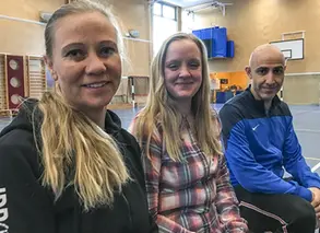 tre lärare sitter på en bänk i gymnastiksalen och tittar in i kameran