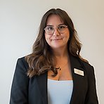 Melisa har långt mörkt hår, glasögon, mörk kavaj och ljusblå topp och ler lätt mot kameran.