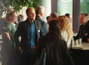 En grupp personer i kostymer och kavajer samlas vid ett bord med kaffemuggar. En man bär en blå skjorta och ett visitkort med texten 