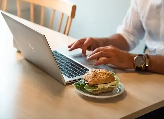 Person arbetar vid laptop vid ett bord med en smörgås på tallrik bredvid.