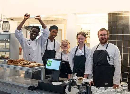 Fem glada elever i restaurangmiljö står bakom disk och presenterar caféutbud.