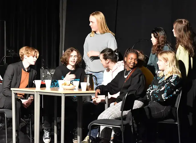 En teaterpjäs med ett gäng ungdomar som sitter och står runt ett fikabord och pratar med varandra.